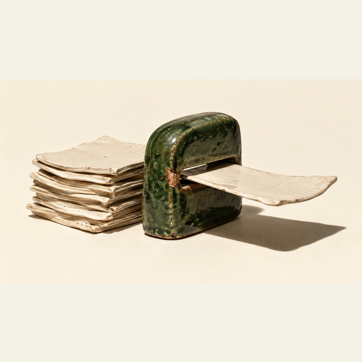 Uneven ivory ceramic sheets stack behind a chipped forest-green ceramic gateway, with one sheet extending through its narrow slot. Hard shadows fall across a warm ivory background.