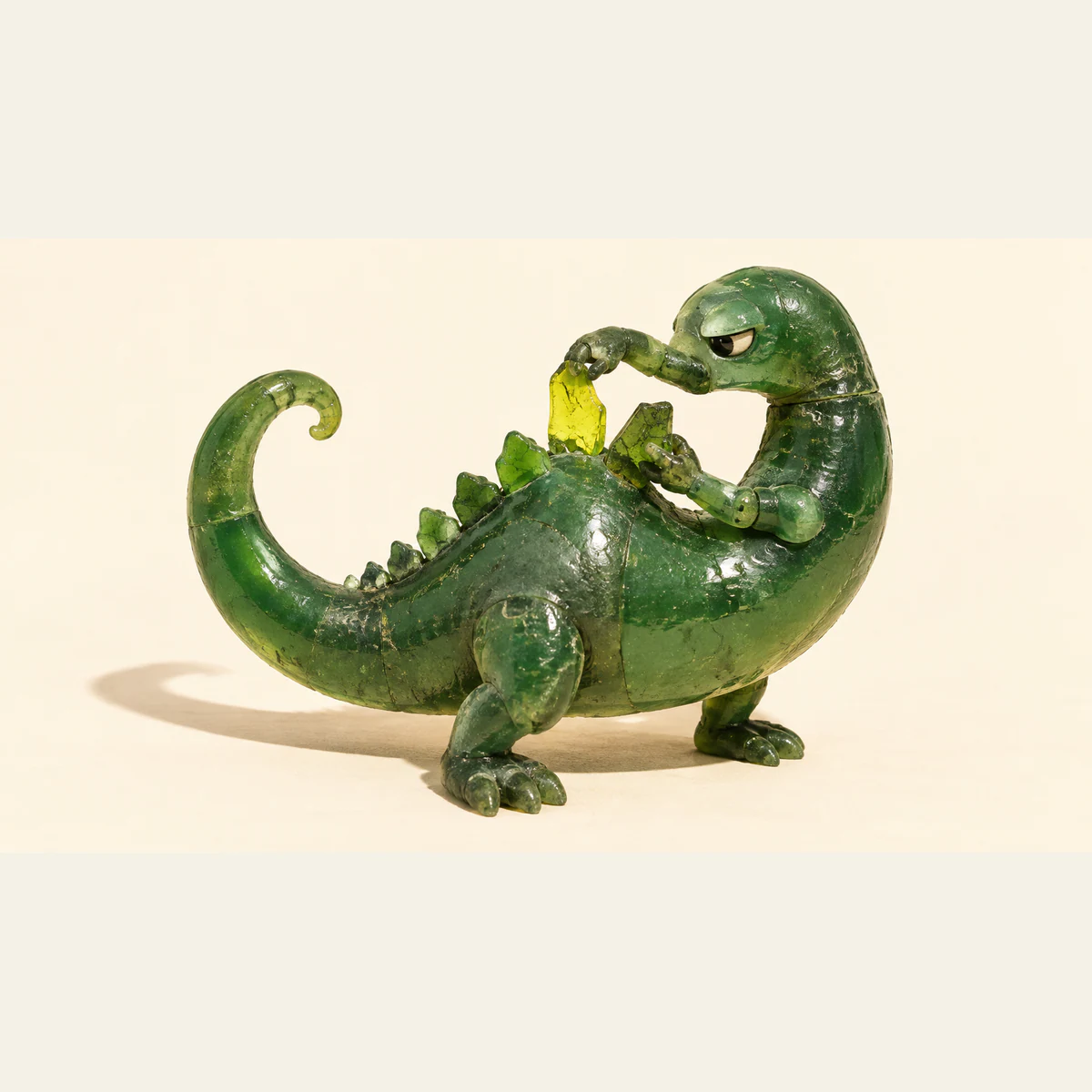 A scuffed translucent green resin dinosaur holds a yellow scale above its back while steadying a tilted green scale, casting a long shadow on warm ivory.