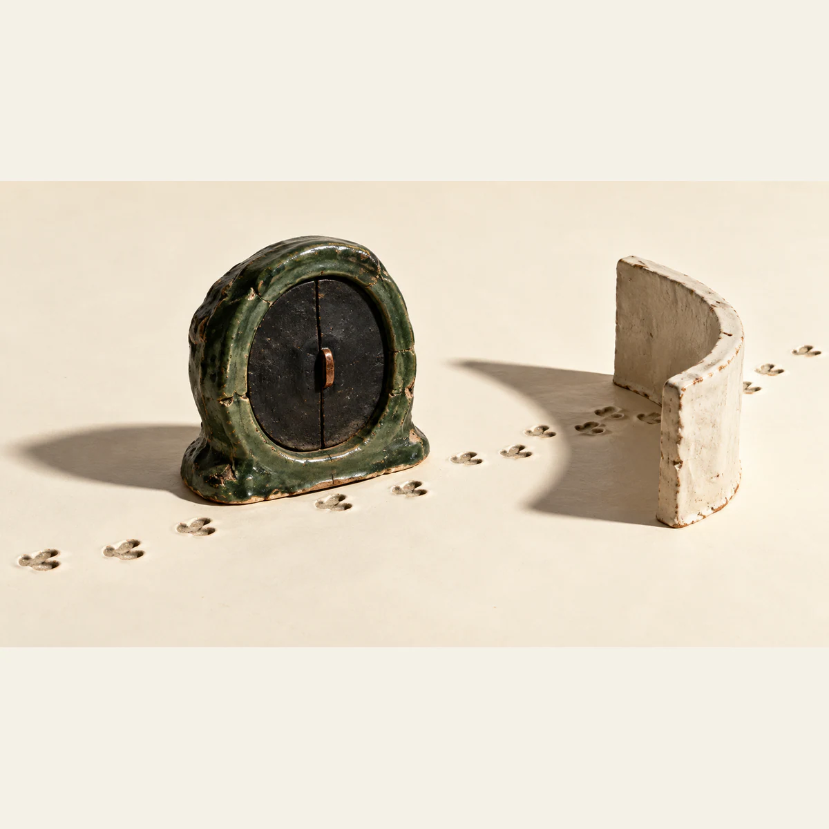 A chipped forest-green ceramic portal with closed black shutters stands beside footprints crossing an ivory surface and passing behind a curved ceramic wall. Hard shadows emphasize the handmade textures.
