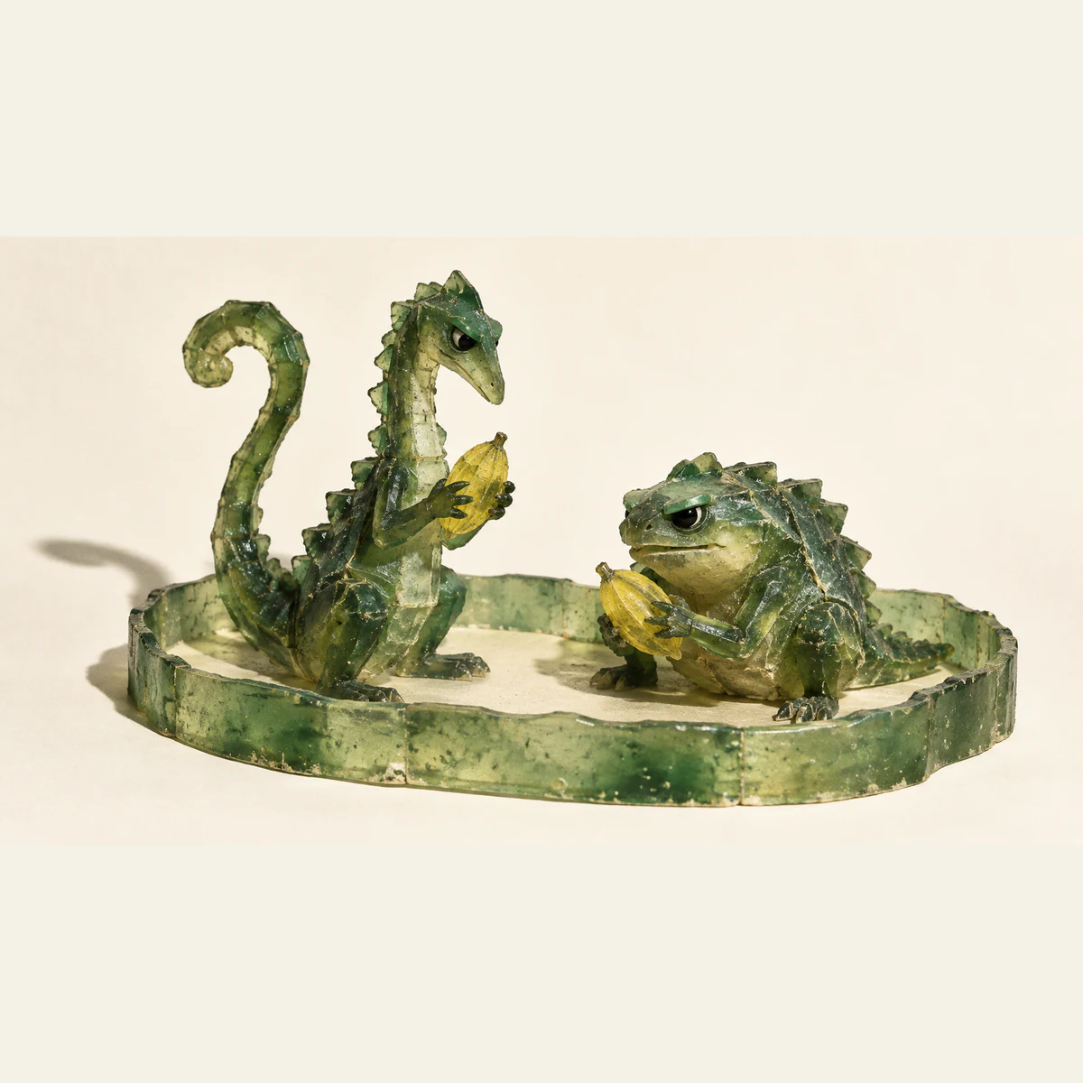 Two scuffed translucent green dinosaur creatures, one tall and one squat, each hold a separate yellow pod inside a low oval resin enclosure on an ivory background.