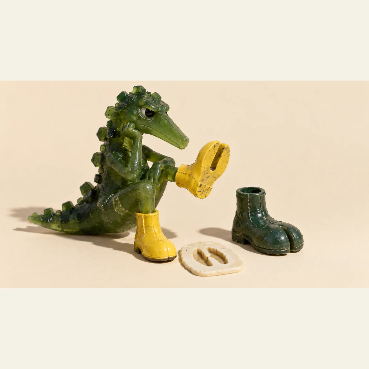 A skeptical translucent green dinosaur raises a scuffed yellow boot beside an unused green boot and an ivory tile bearing a split footprint. Hard shadows fall across a warm ivory background.