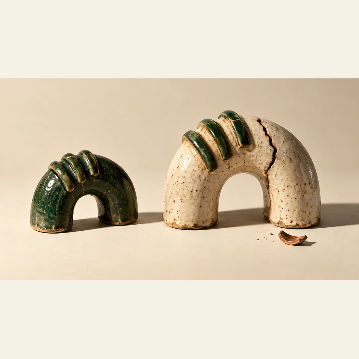 Two handmade ceramic arches share three raised green ridges. The smaller green arch is intact; the larger ivory arch has a deep crack, with a copper-colored shard below. Hard shadows cross the warm ivory ground.