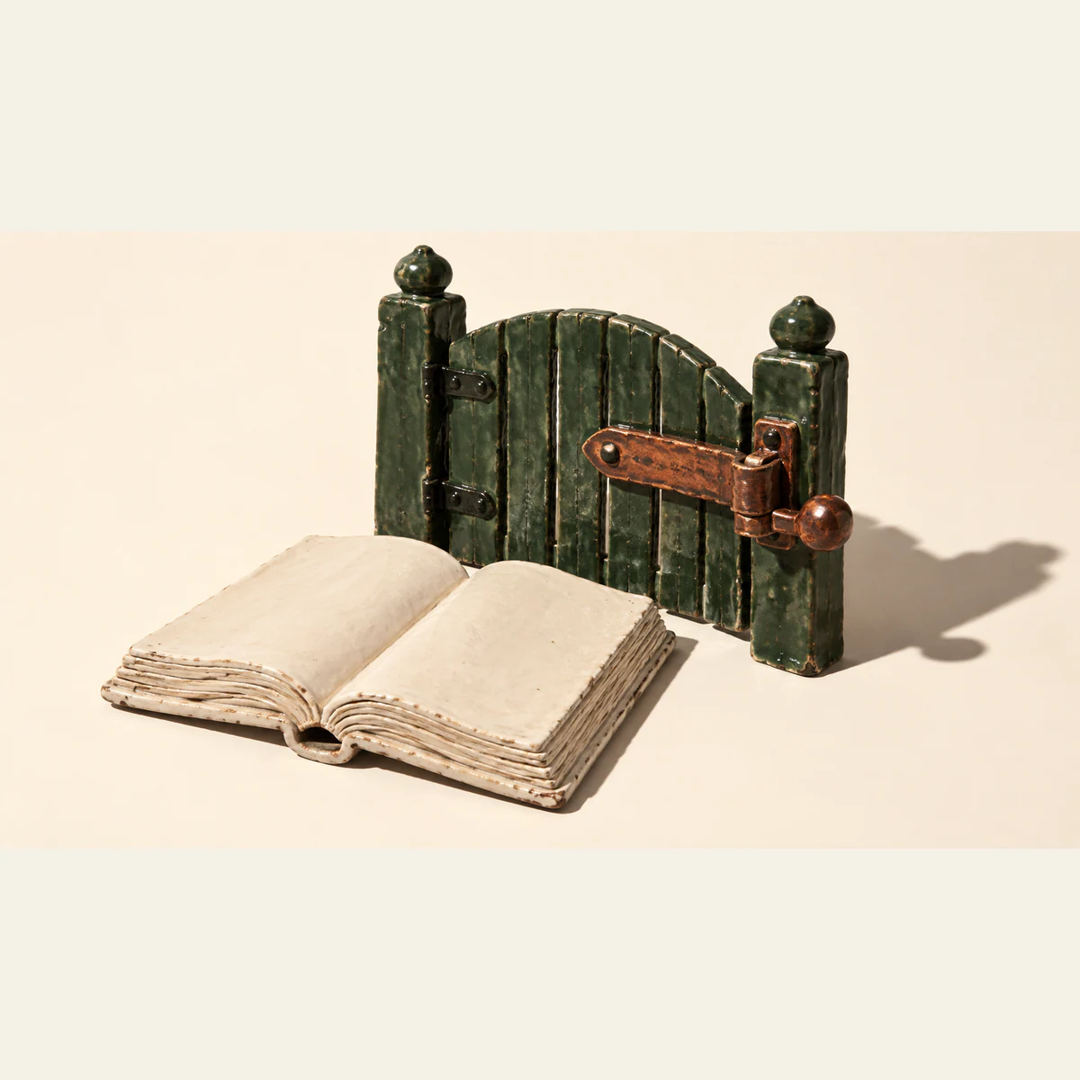 An open, blank ceramic book sits before a closed forest-green miniature gate with an oversized copper-colored latch, casting hard shadows across an ivory background.