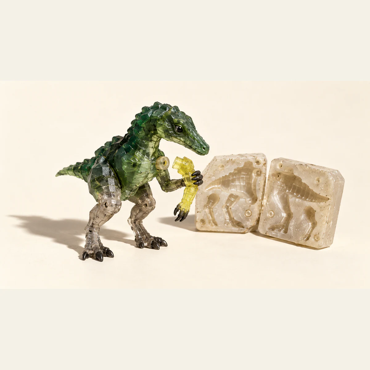 A translucent green resin dinosaur examines a detached yellow arm beside an open casting mold. Visible joints, bubbles and scuffs catch hard light against an ivory background.