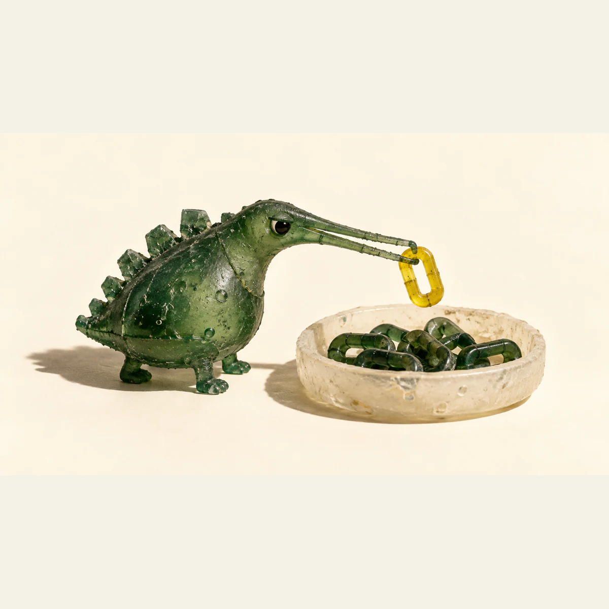 A bubble-speckled green resin dinosaur with a skeptical eye lifts a yellow link with its tweezer-like snout above an ivory dish of green links, casting hard shadows on a warm ivory background.