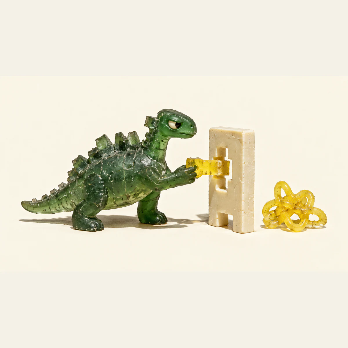 A skeptical translucent green resin dinosaur pushes a yellow keyed peg into an ivory gate. A separate yellow tangle rests beside it, with hard shadows on a warm ivory background.
