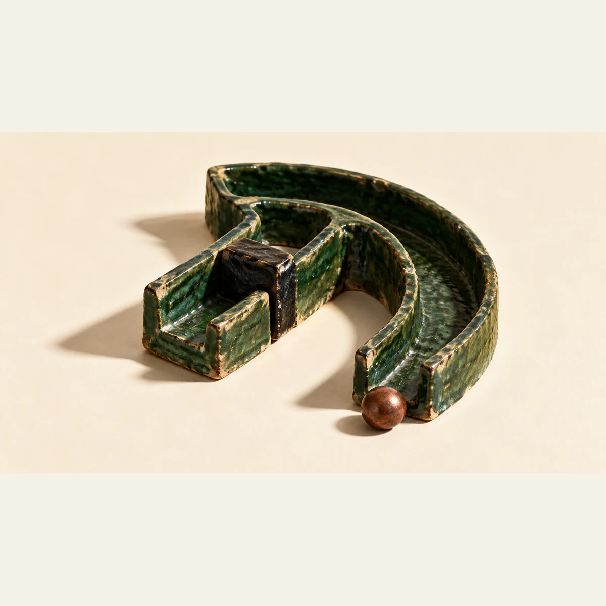 A chipped forest-green ceramic channel forks into a passage blocked by a black stopper and an open curved route. A copper-colored bead rests at the open outlet on an ivory surface with strong shadows.