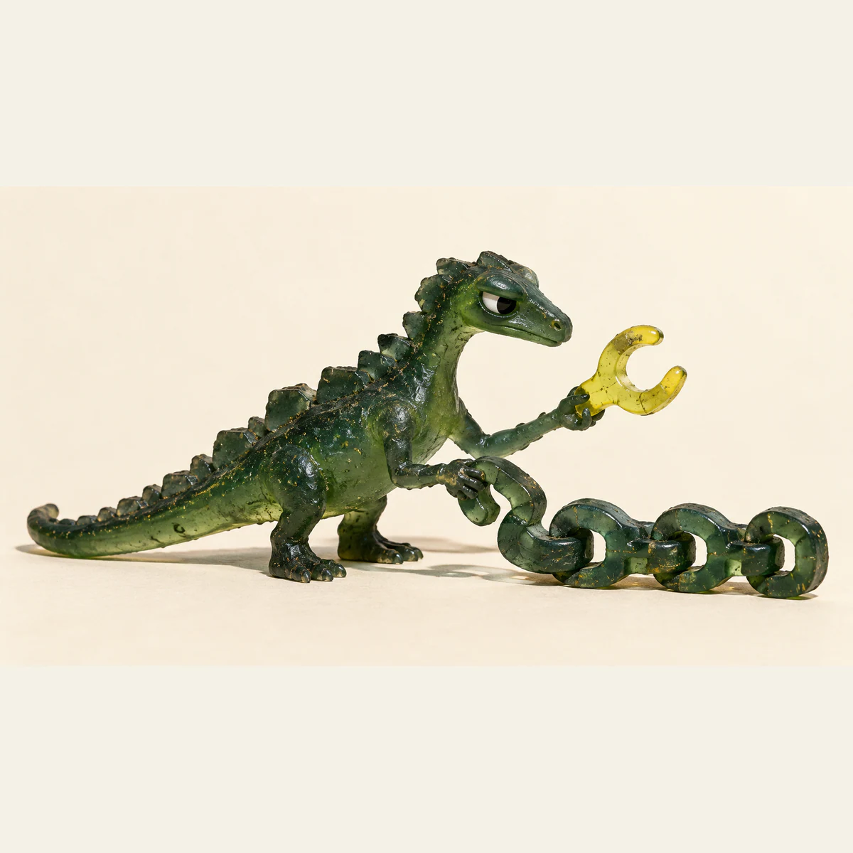 A skeptical translucent green resin dinosaur grips a short green chain while holding a yellow forked piece apart, on a warm ivory background with hard shadows.