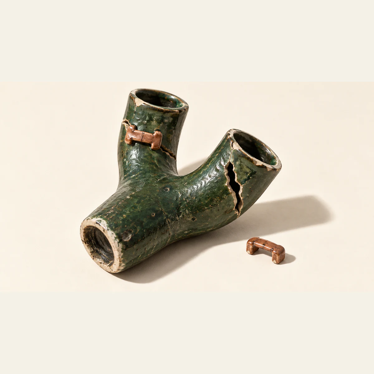 A chipped forest-green ceramic Y-shaped vessel on warm ivory. A copper-colored staple bridges one branch’s crack; the other branch remains split, with a loose staple beside it. Hard light casts a long shadow.