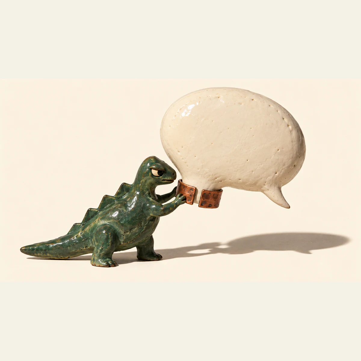 A skeptical green ceramic dinosaur holds an open copper-colored collar around the narrow base of an oversized blank ivory speech bubble. Handmade surface imperfections and long shadows stand out against a warm ivory background.