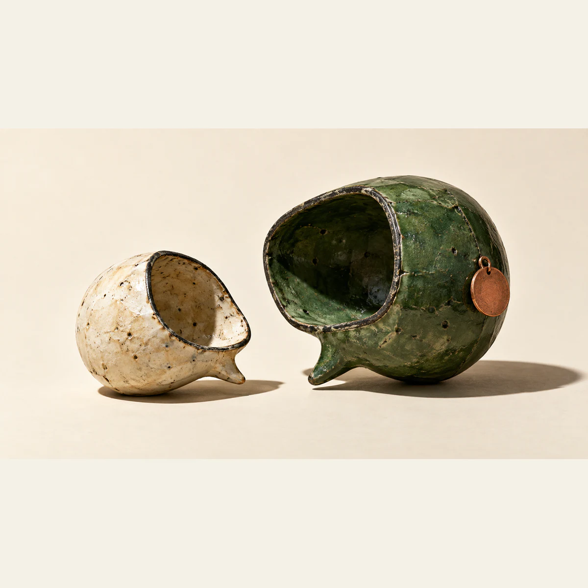 Two hollow, weathered ceramic speech bubbles face each other on an ivory surface. The larger forest-green form carries a blank copper-colored tag; the smaller form is cream.