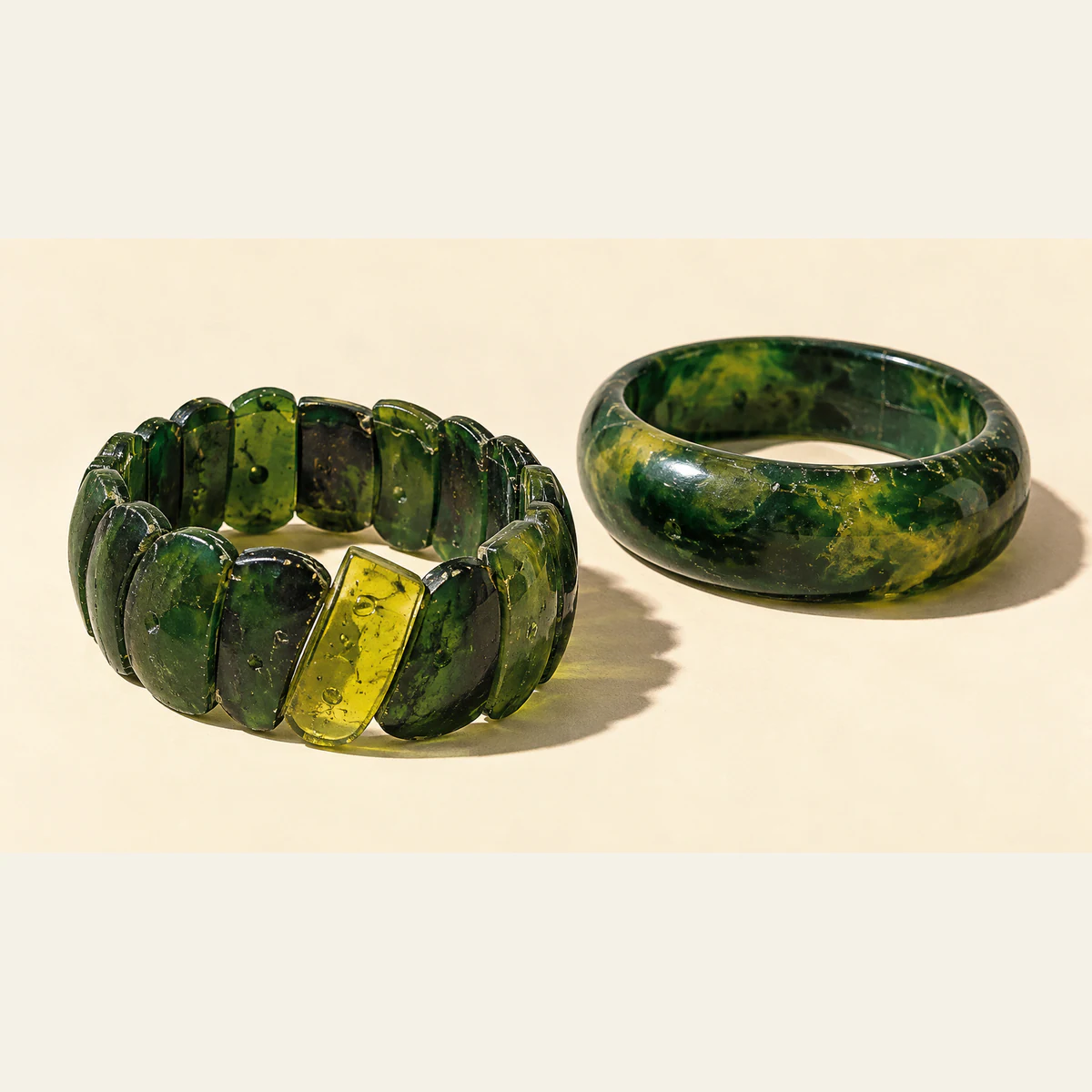 Two imperfect green resin bracelets cast hard shadows on ivory. The segmented bracelet has one tilted yellow piece; the smooth bangle contains diffuse yellow mottling.
