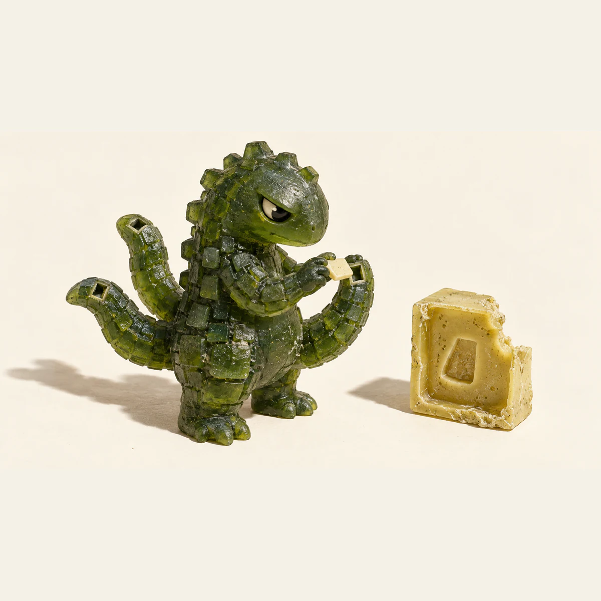 A skeptical translucent green resin dinosaur holds an ivory scale beside a socket in one of three curling tails. A rough yellow mold with a missing corner stands nearby on an ivory surface, casting a hard shadow.