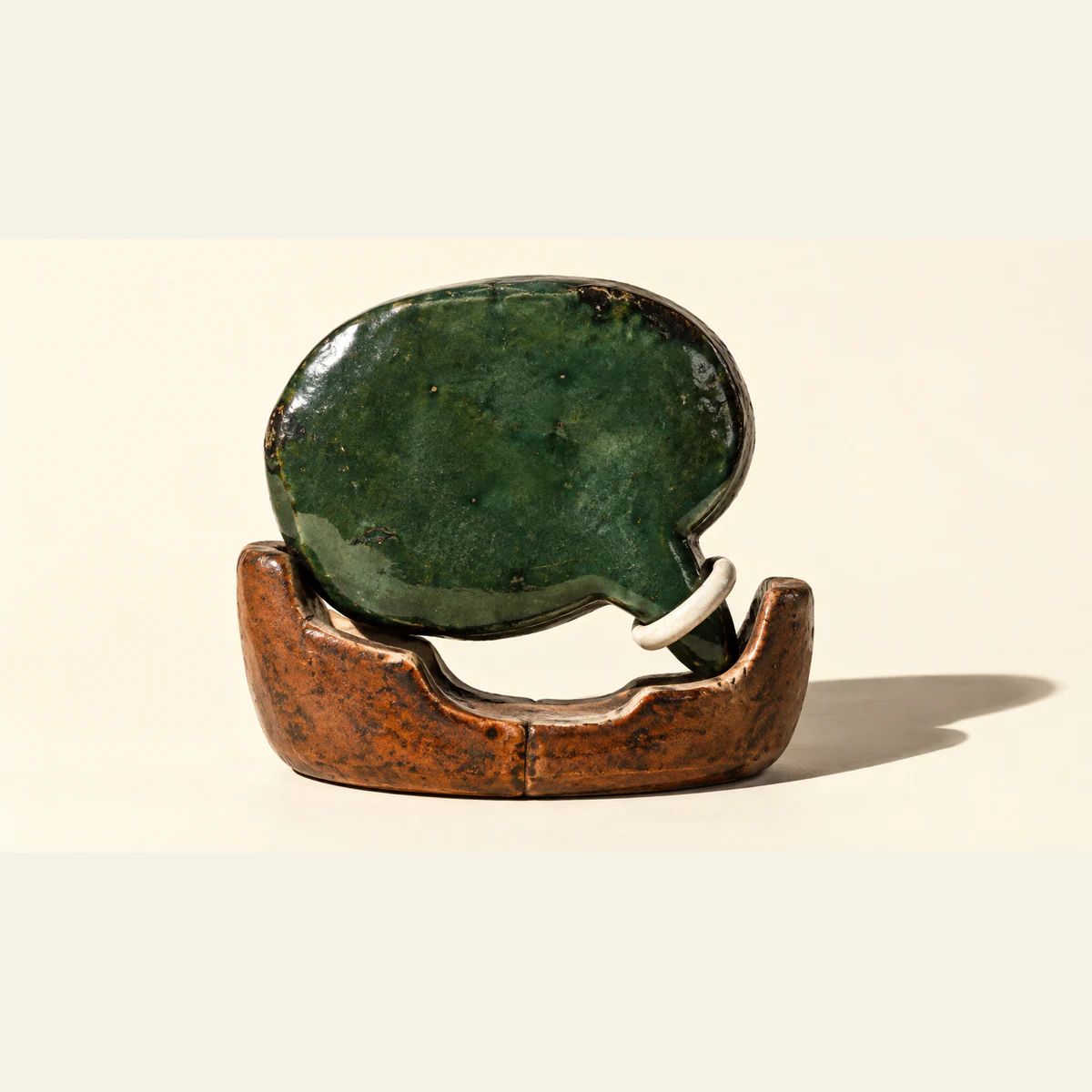 A worn forest-green ceramic speech bubble rests unevenly in a copper-colored cradle, leaving a broad gap. An ivory ring encircles its tail. Hard shadows fall across the warm ivory background.