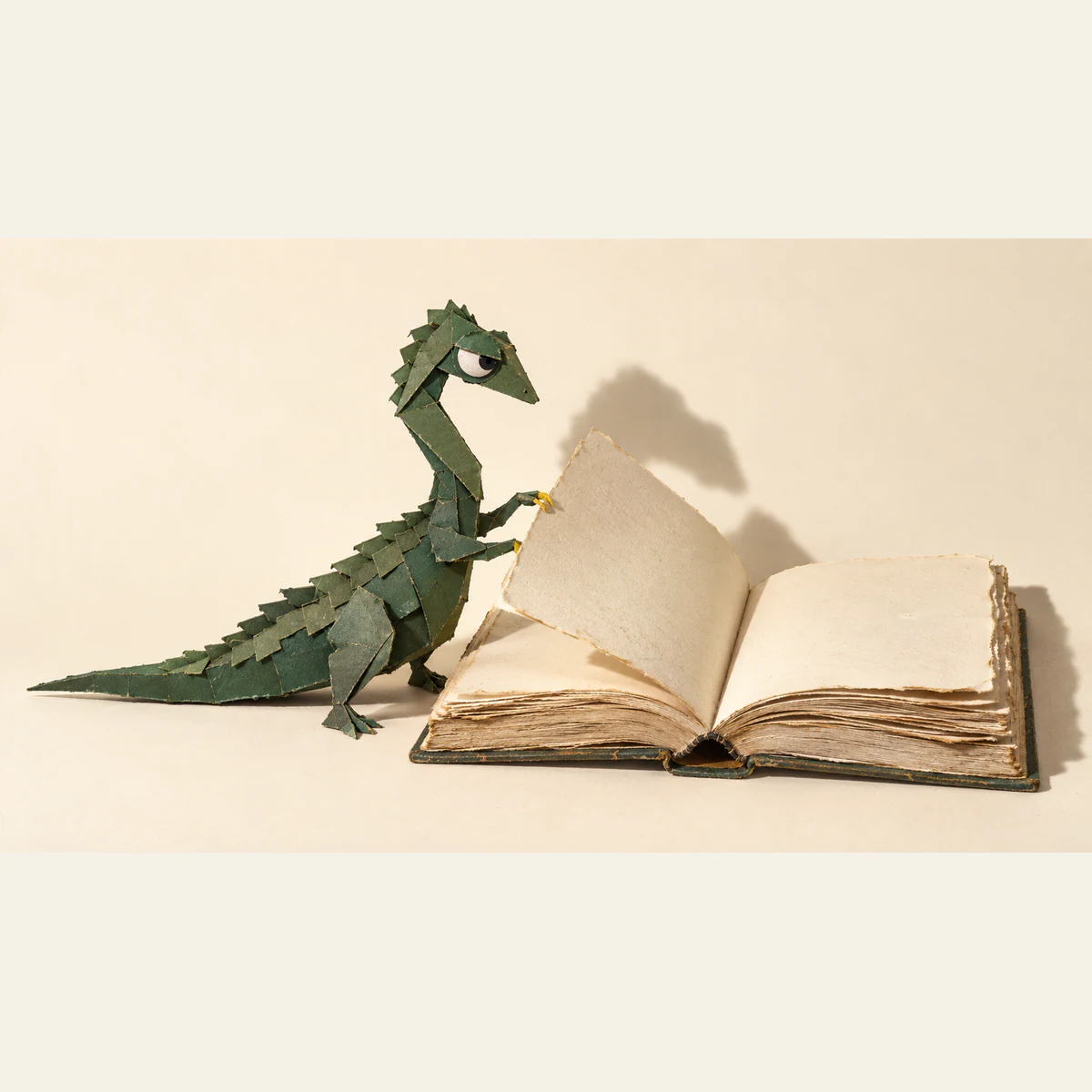 A forest-green paper dinosaur with a skeptical eye and yellow claws lifts a blank, rough-edged page of an oversized open book. Handmade seams and strong shadows stand out against warm ivory.