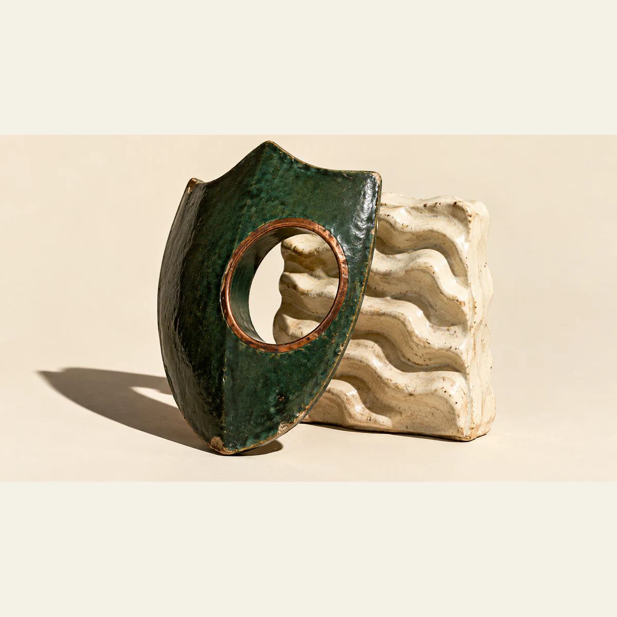 A scuffed forest-green ceramic shield with a copper-rimmed circular opening leans before a ridged ivory ceramic slab, casting a long shadow across a warm ivory background.