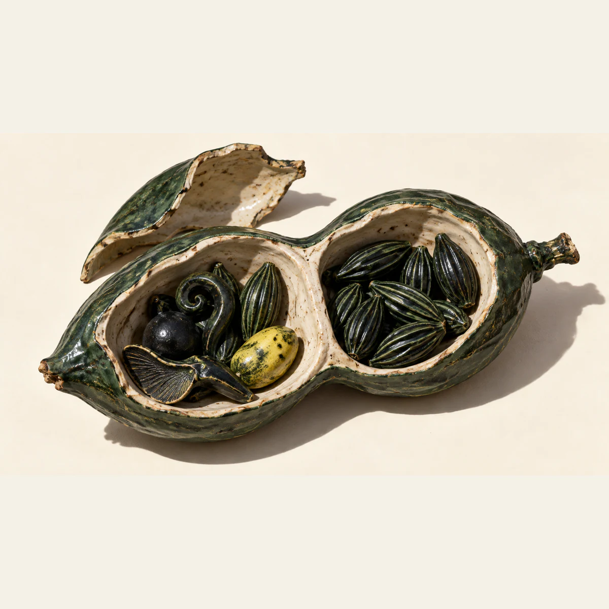 An opened green ceramic seedpod on ivory holds varied sculptural seeds in its left chamber and matching ridged seeds in its right. A detached shell rests behind it, casting a hard shadow.