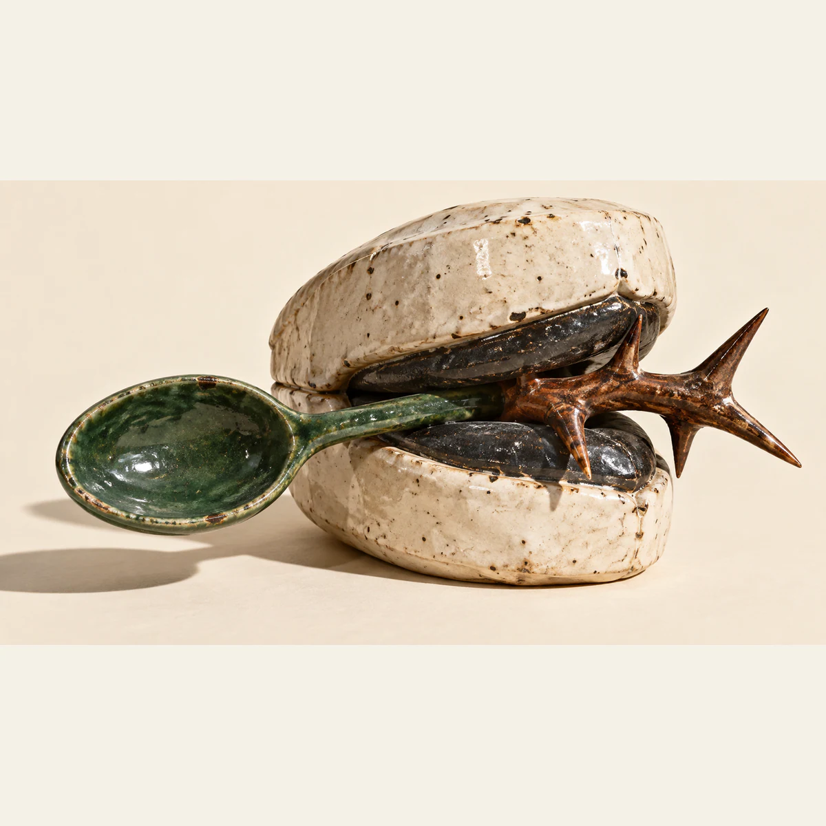 An ivory ceramic clasp with black inner jaws traps a green spoon beside a copper-colored thorn branch, casting hard shadows across a warm ivory surface.