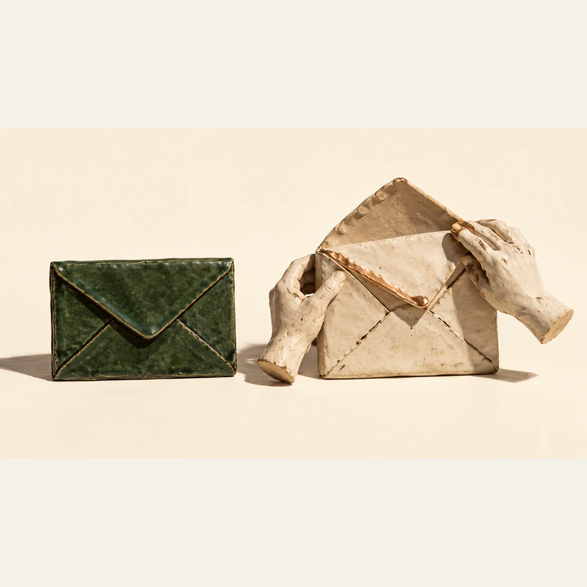 A closed forest-green ceramic envelope sits beside a larger open ivory envelope held by two sculpted ceramic hands. Copper accents, rough seams and hard shadows stand out against a warm ivory background.