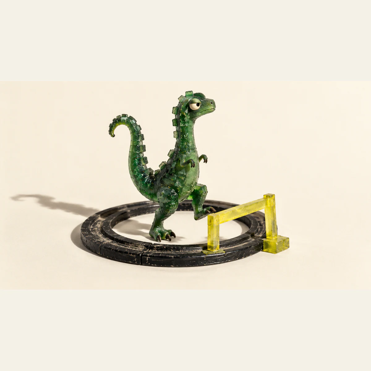 A skeptical green resin dinosaur raises one foot beside a yellow barrier on a scuffed black circular track, casting a long shadow across an ivory background.