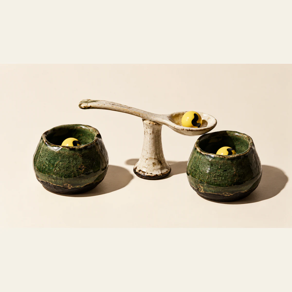 Two rough forest-green ceramic vessels contain yellow pellets with black markings. Between them, an ivory ceramic spoon on a pedestal holds a matching pellet above the right vessel, casting hard shadows on a warm-ivory background.