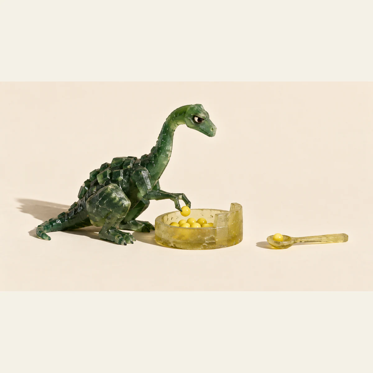A skeptical green resin dinosaur lifts a yellow pellet from a shallow dish. A separate resin spoon holds one pellet on the ivory ground, with hard shadows beneath the handmade objects.