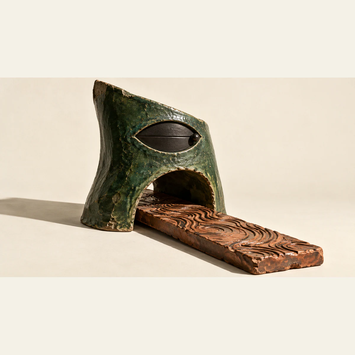 A weathered forest-green ceramic hood has a closed black eye-shaped shutter above an open arch. A grooved copper-colored slab passes through the arch, casting a hard shadow on warm ivory.