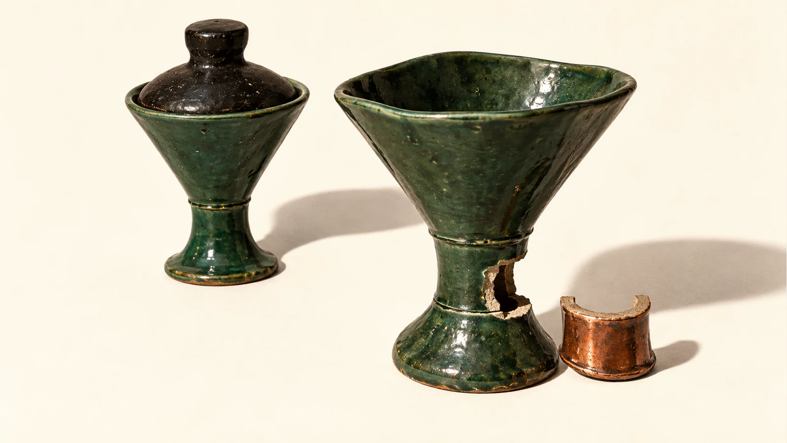 Two forest-green ceramic funnels cast hard shadows on ivory. One has a black stopper; the other has a gap in its stem beside a copper-colored replacement segment.