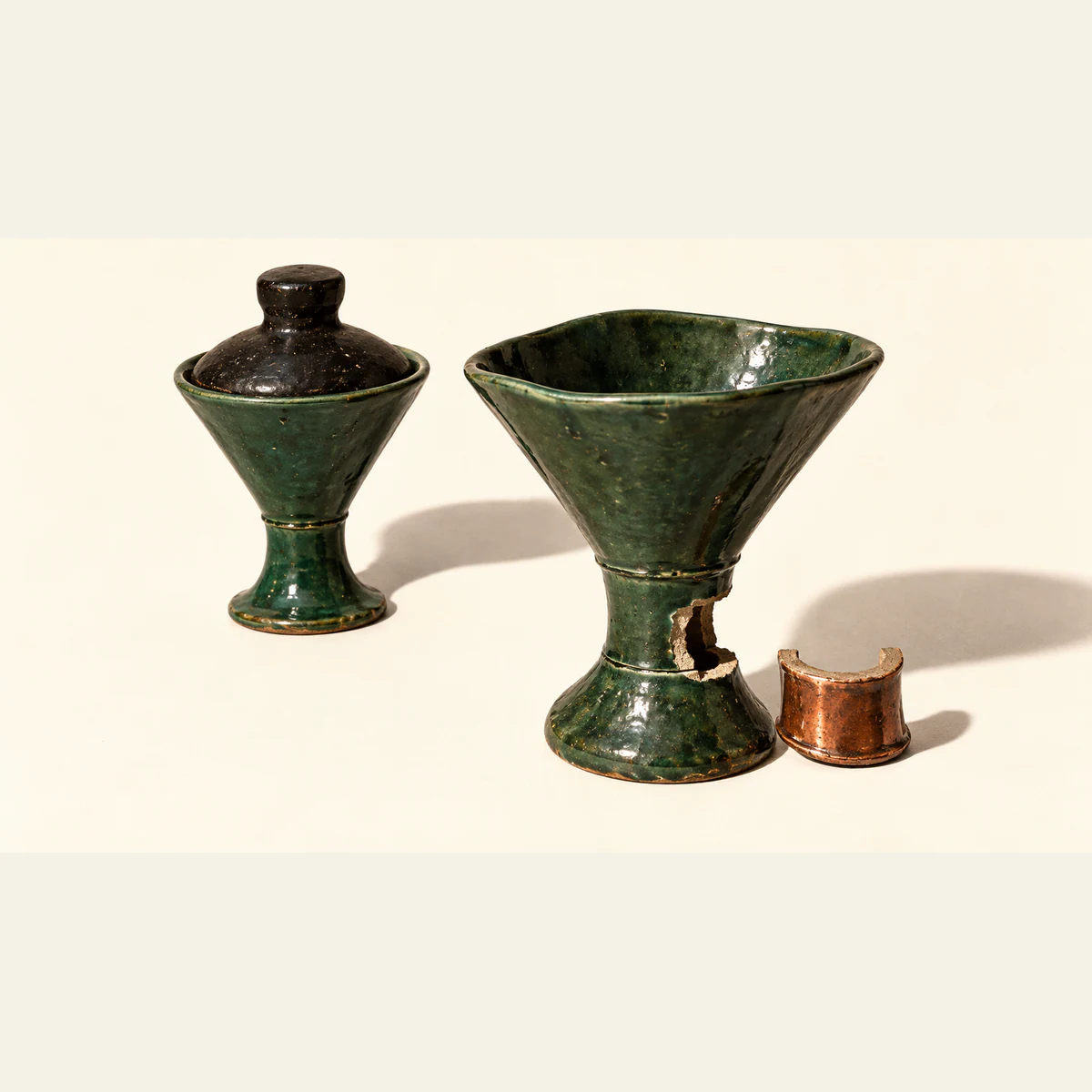 Two forest-green ceramic funnels cast hard shadows on ivory. One has a black stopper; the other has a gap in its stem beside a copper-colored replacement segment.