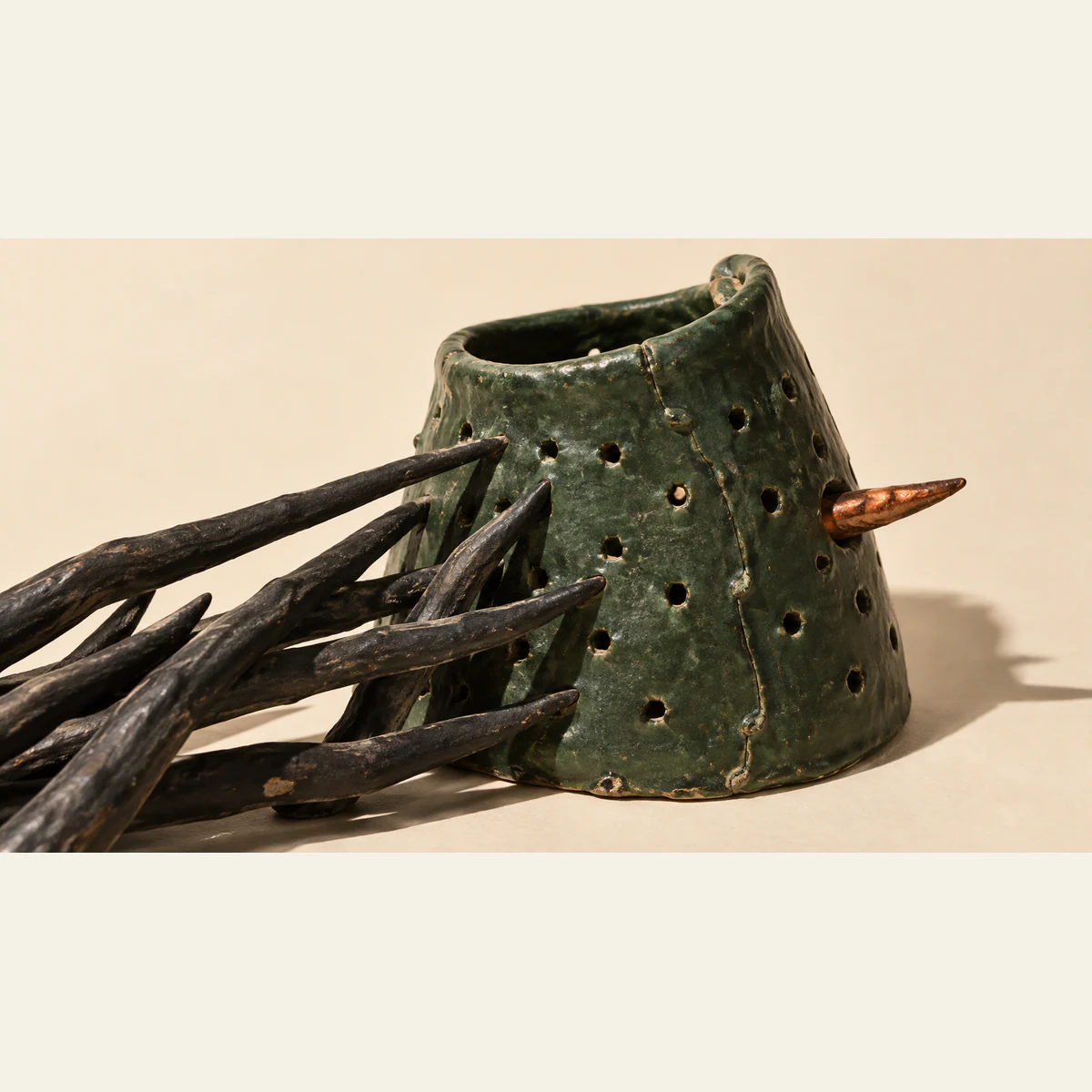 Black ceramic thorns press against a weathered, perforated green ceramic collar, with a copper-colored point protruding from its right side. Hard shadows fall across an ivory background.