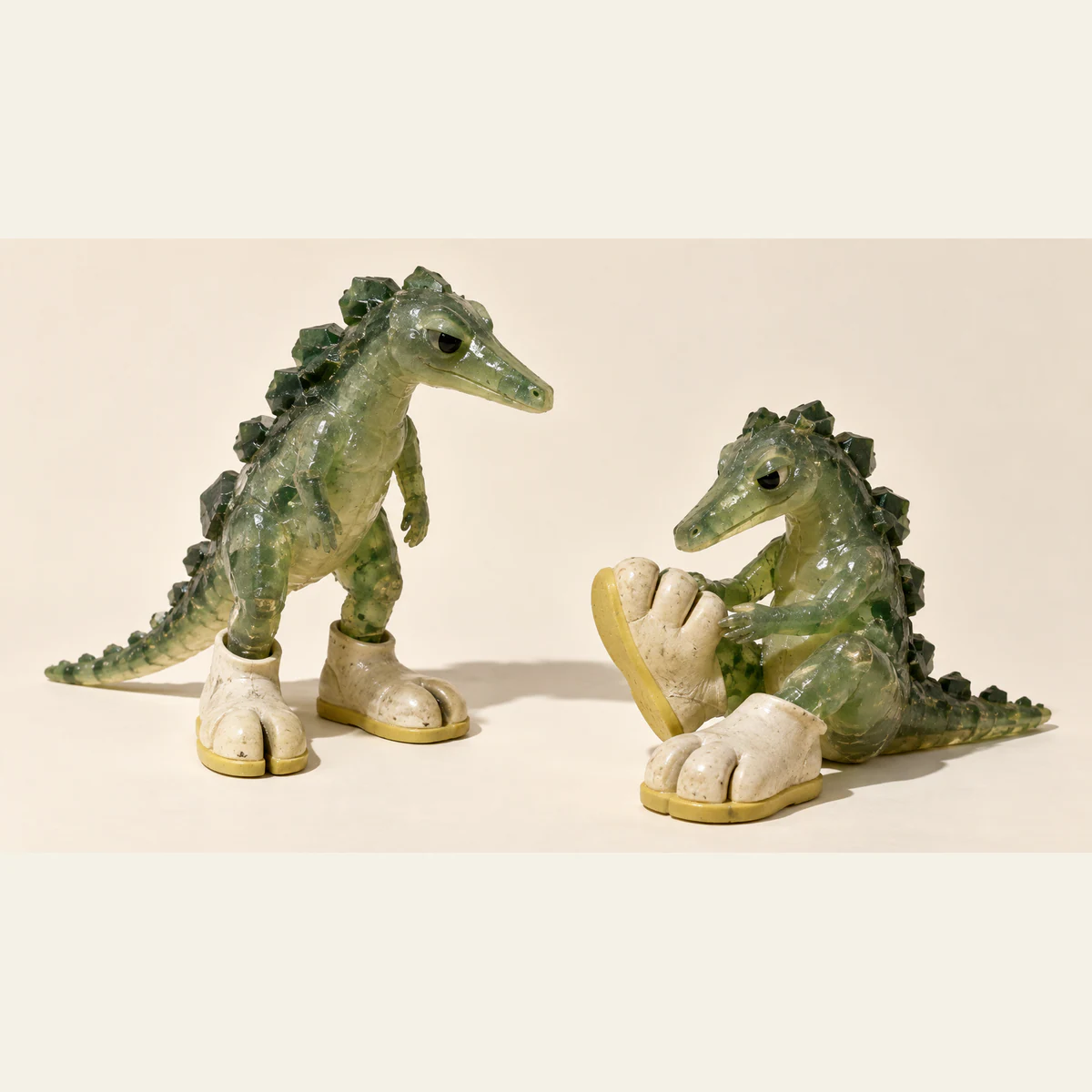 Two translucent green resin dinosaurs wear oversized ivory shoes with yellow soles. One stands in split-toe boots; the other sits inspecting a three-toed shoe against a warm ivory background.
