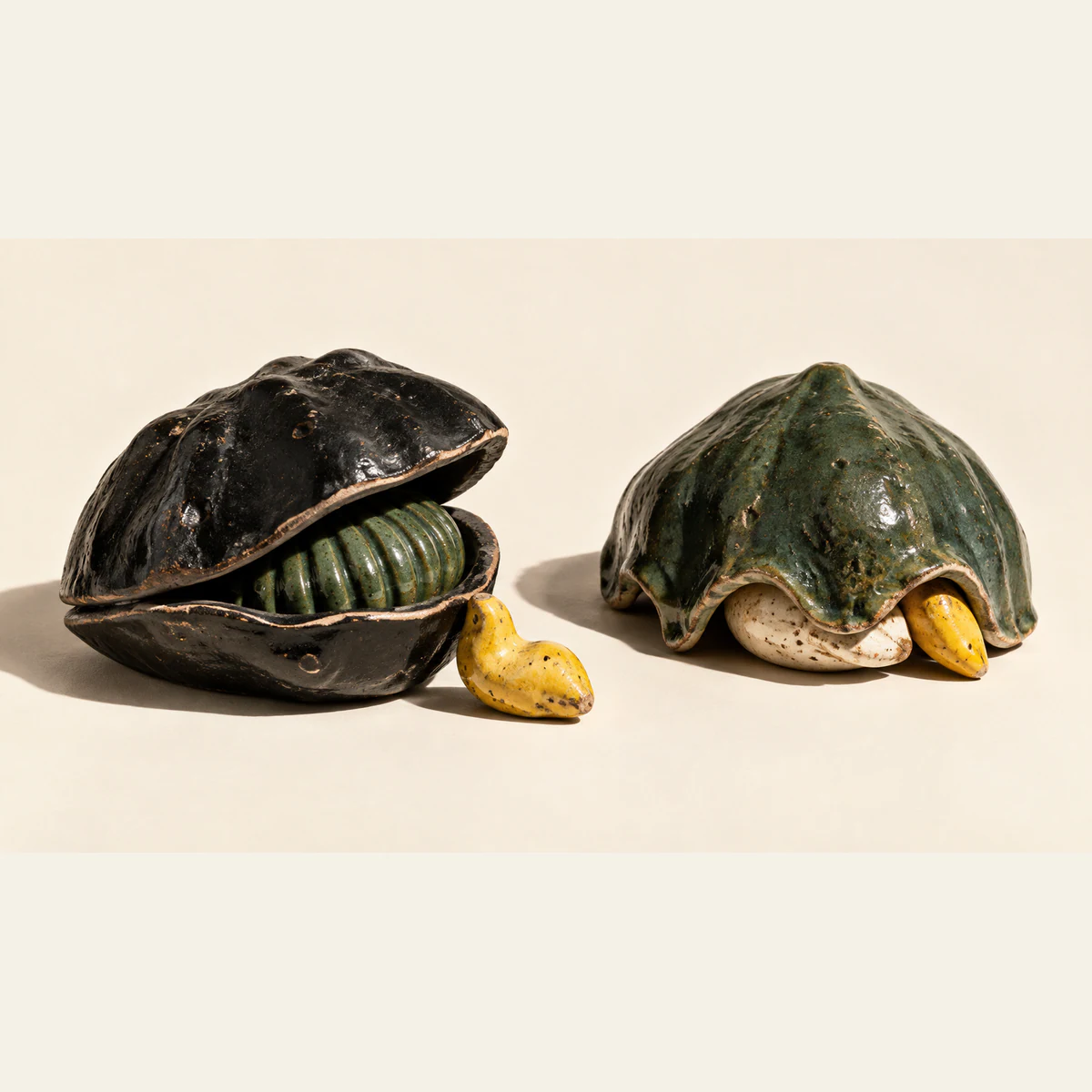 Two chipped ceramic shelters on ivory: a black clamshell holds a ribbed green form beside an uncovered yellow piece; a green canopy partly covers ivory and yellow forms.