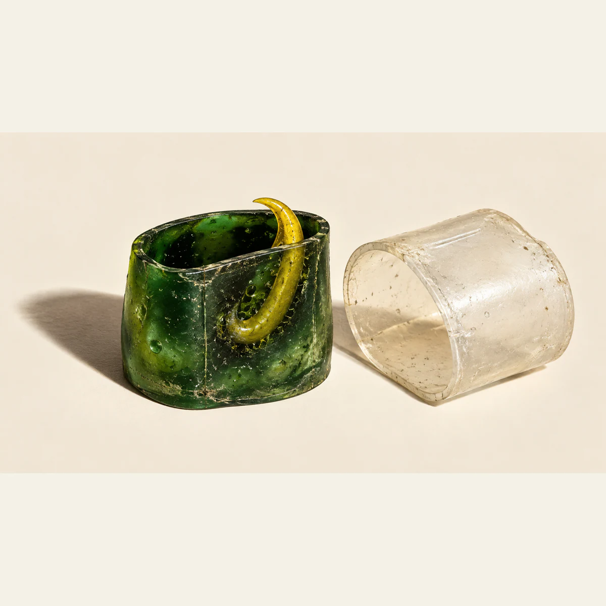 A yellow hooked barb remains embedded inside a scuffed green resin vessel beside a detached clear sleeve, casting hard shadows across an ivory surface.