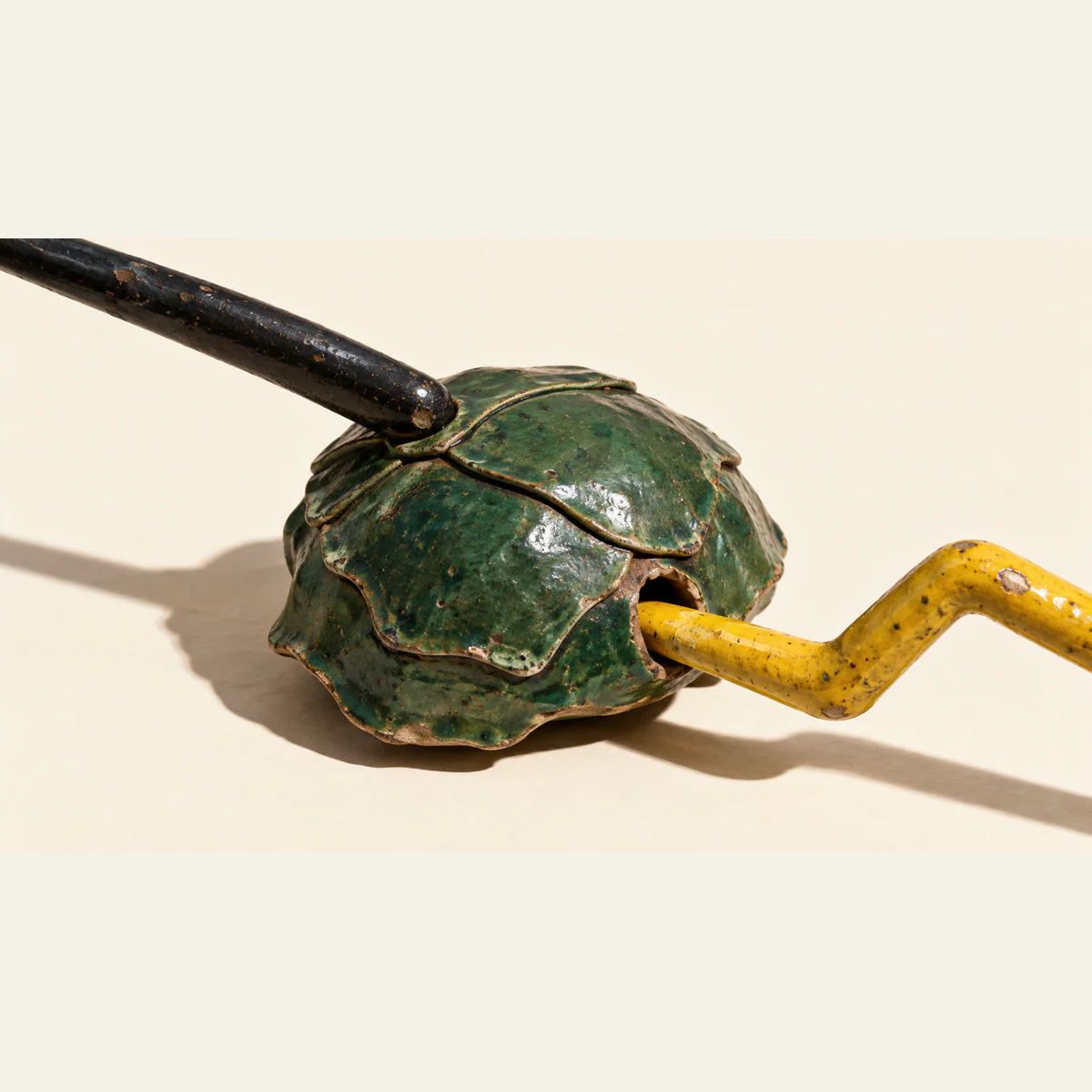 A chipped green ceramic shell on ivory, with a black probe touching its overlapping plates and a bent yellow probe entering a side opening. Hard shadows extend across the background.