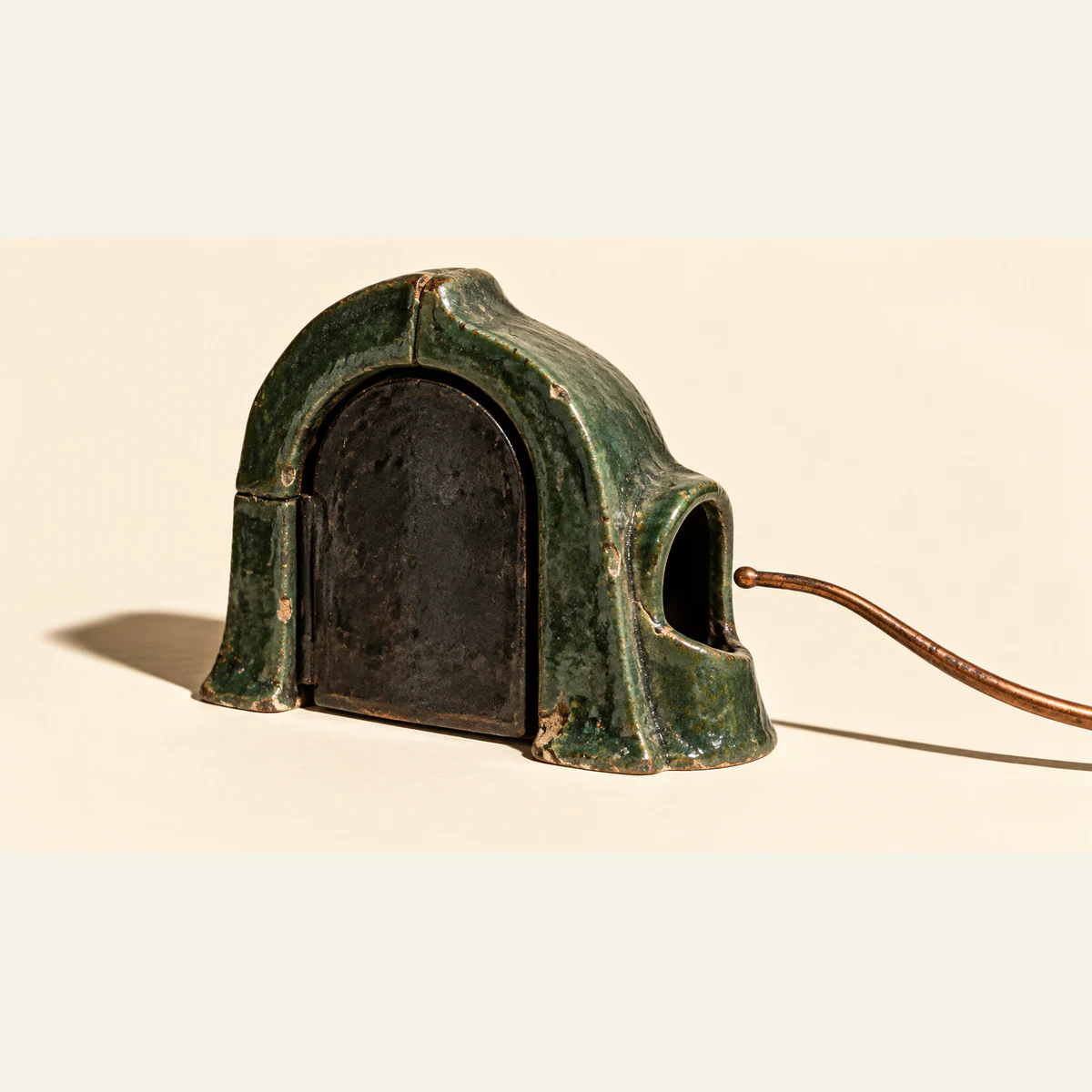 A chipped forest-green ceramic enclosure has a closed black front door and a dark side opening. A copper-colored probe curves in from the right, stopping outside the opening, against warm ivory with hard shadows.