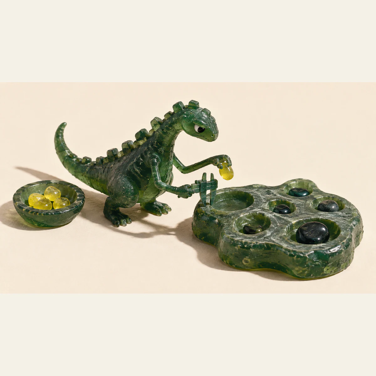 A skeptical green resin dinosaur holds a yellow pebble above a hollow while pressing calipers against its rim. A separate bowl holds yellow pebbles; other hollows contain dark stones. Hard shadows fall across an ivory background.