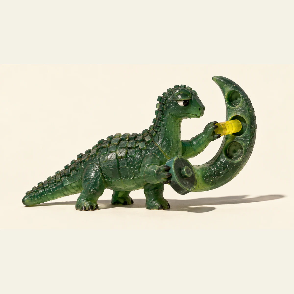 A skeptical green resin dinosaur inserts a yellow peg into the middle of three sockets on a curved appendage while holding a detached wheel, casting a hard shadow across an ivory background.