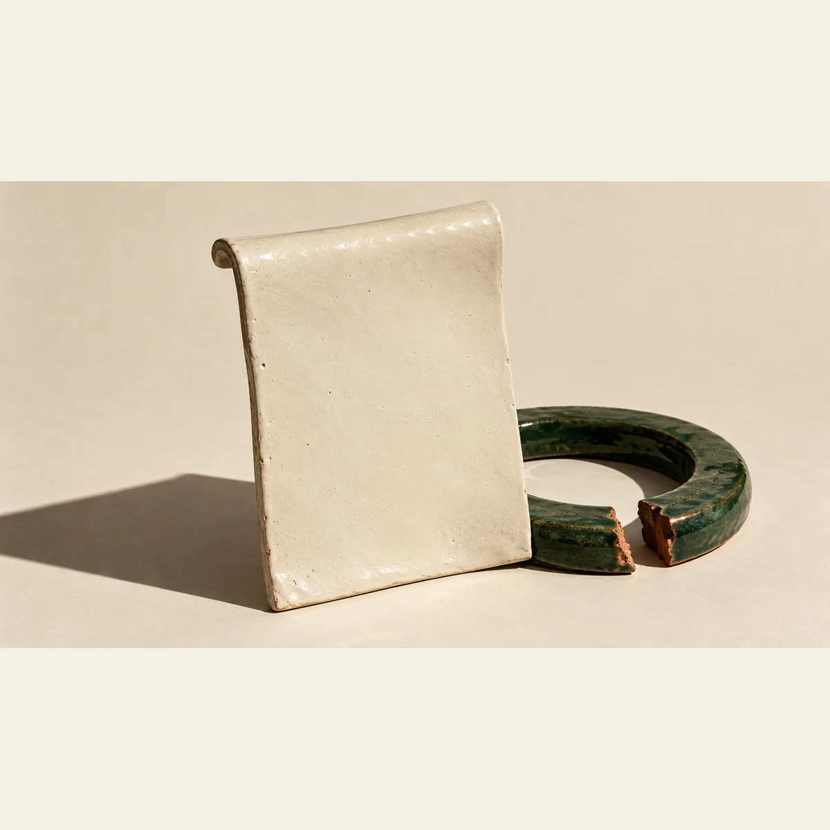 A blank ivory ceramic sheet stands before a broken forest-green ceramic ring, with copper-colored fracture edges and hard shadows on a warm ivory surface.