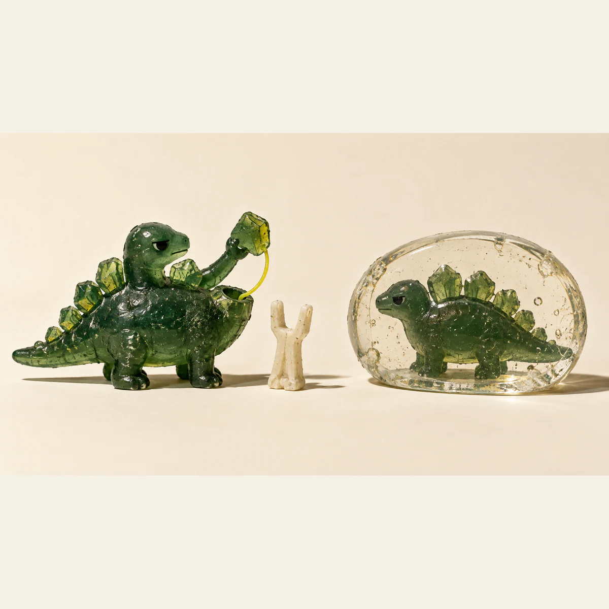 A scuffed green resin dinosaur lifts a detachable piece tethered by a yellow loop to an open socket. An ivory fork-shaped object stands beside it; a second dinosaur sits sealed inside clear resin on the warm ivory background.