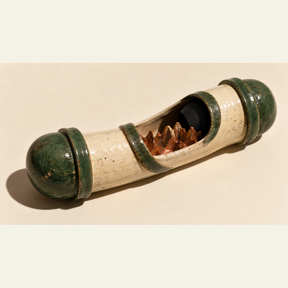 An elongated ivory ceramic vessel with forest-green capped ends lies diagonally on a warm ivory surface. Its open middle reveals copper-colored jagged ridges, while a black hood conceals part of the interior. Worn glaze and a hard shadow emphasize its handmade construction.