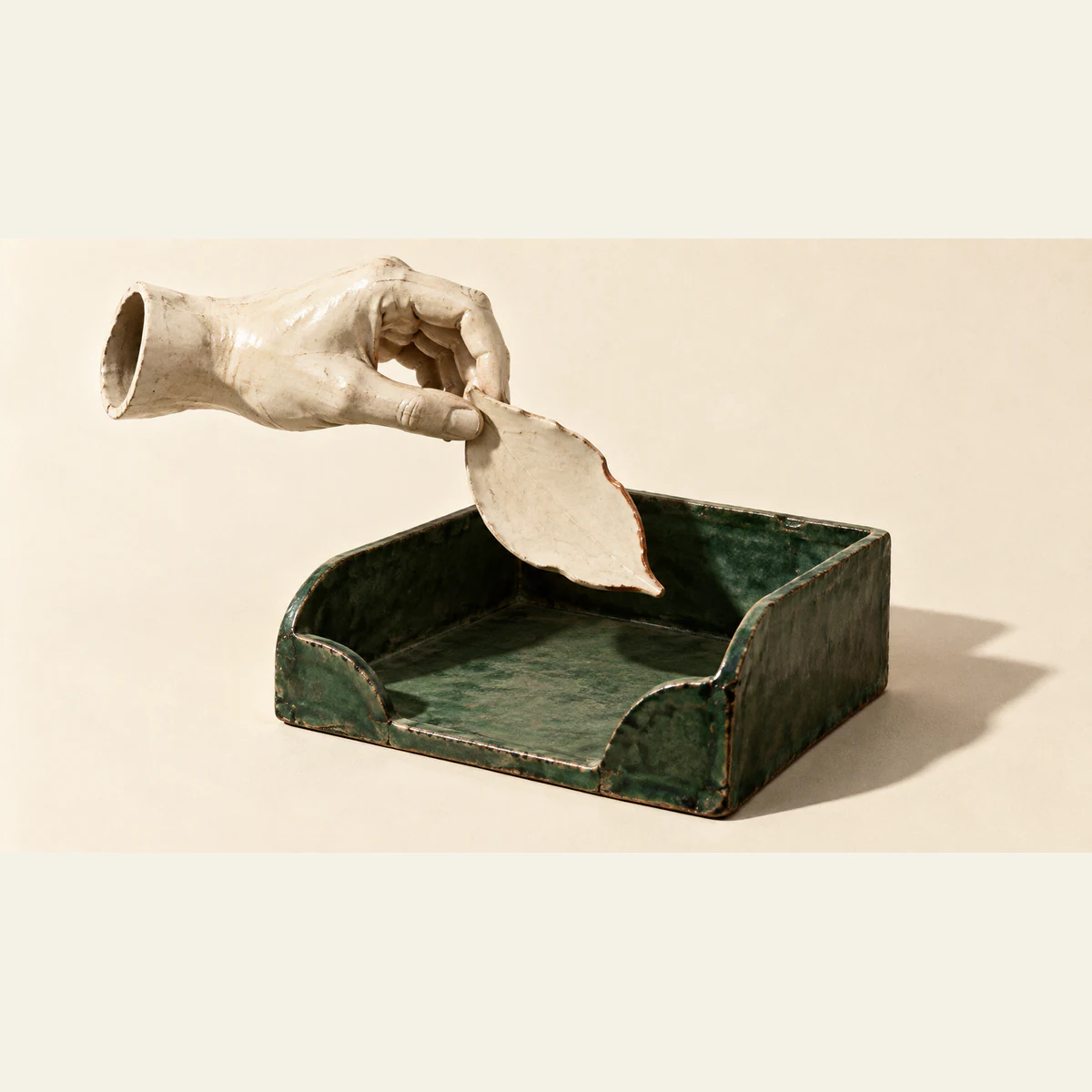 A hollow ceramic hand suspends an irregular ivory leaf with a copper-colored edge above an empty forest-green document tray, casting hard shadows across a warm ivory background.