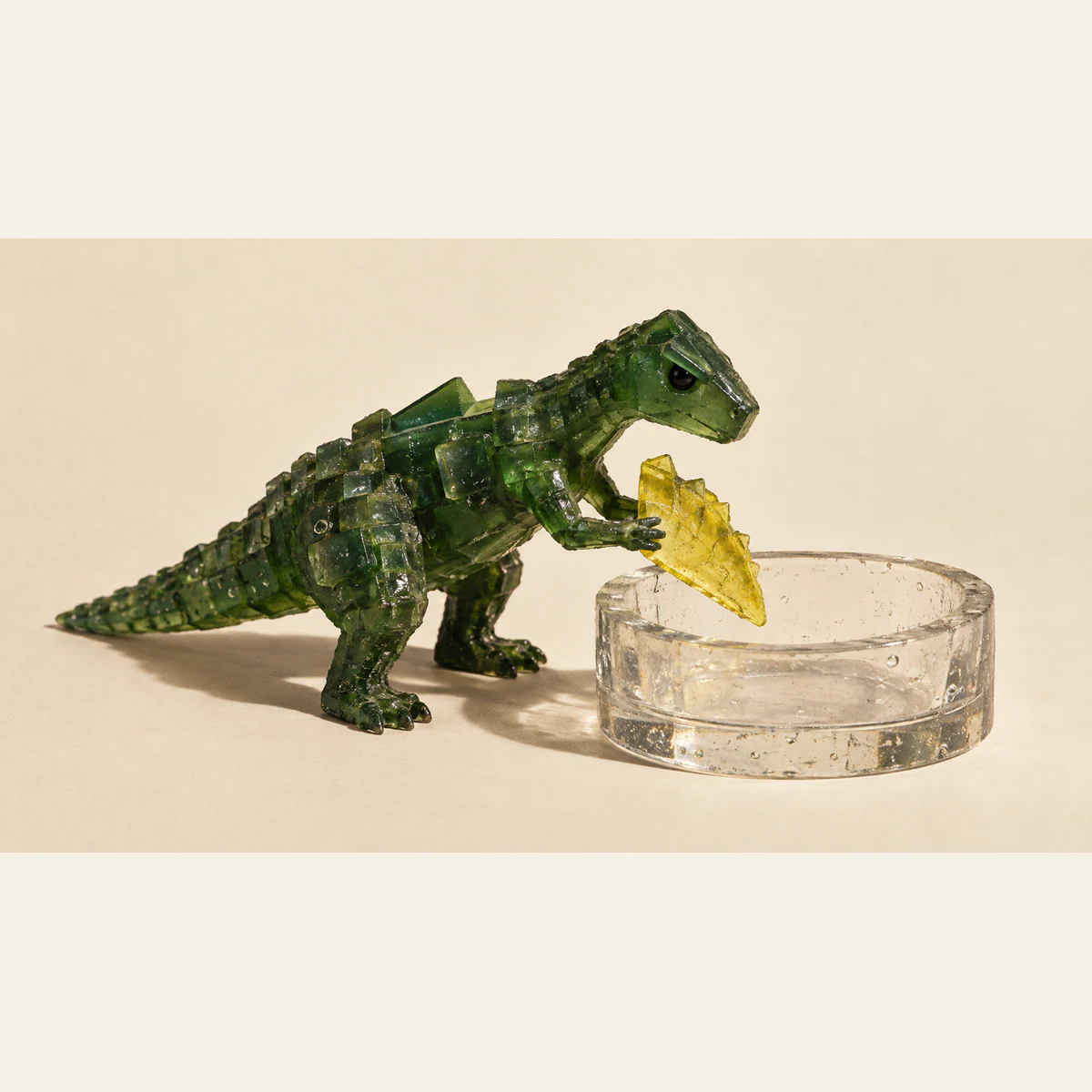 A translucent forest-green resin dinosaur holds a detached yellow fin over a clear, bubble-filled resin basin, casting hard shadows on an ivory background.