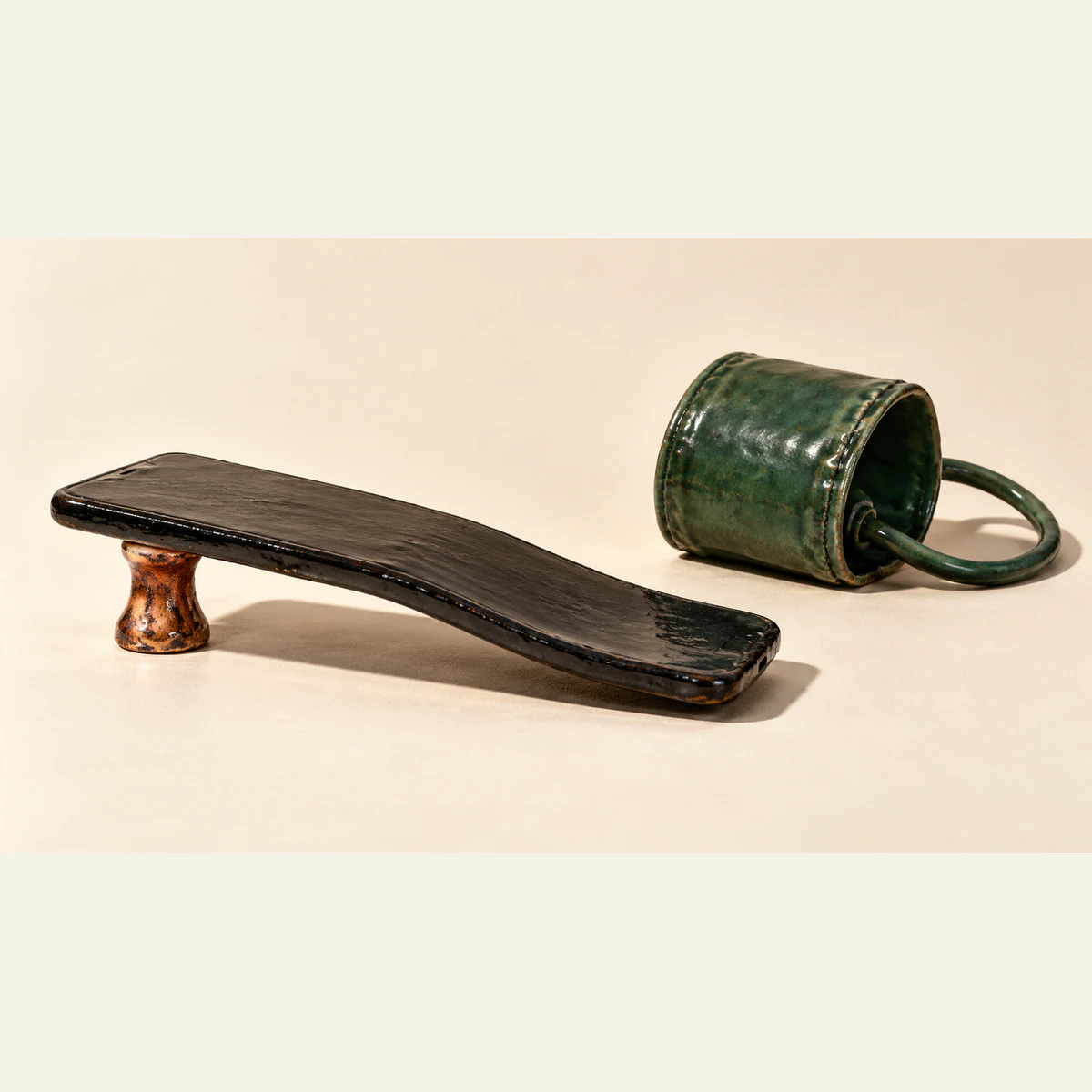 A warped black ceramic smartphone rests on one copper-colored pedestal, its far end drooping toward an ivory surface. A separate green ceramic blood-pressure cuff sits behind it, casting a hard shadow.