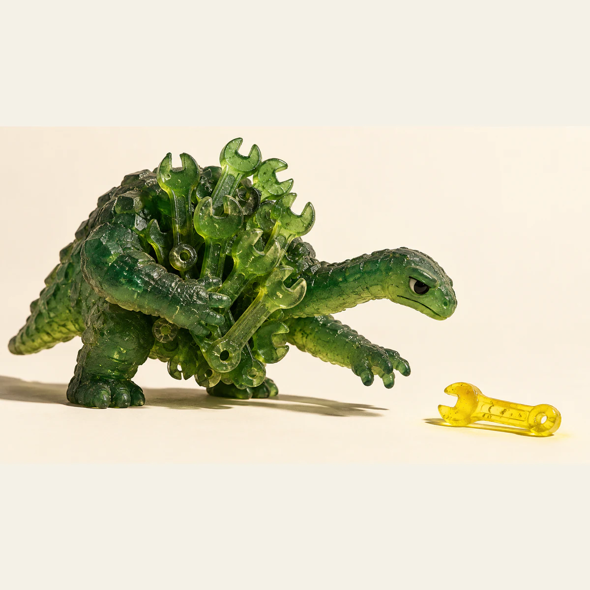 A translucent green resin dinosaur clutches a crowded bundle of green wrenches while reaching toward a solitary yellow wrench on an ivory surface, with hard shadows and visible resin bubbles.