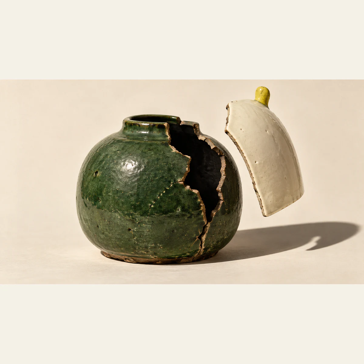 A cracked forest-green ceramic vessel beside a lifted ivory shell with a yellow tab. Its jagged opening exposes a dark interior, casting hard shadows across a warm ivory background.