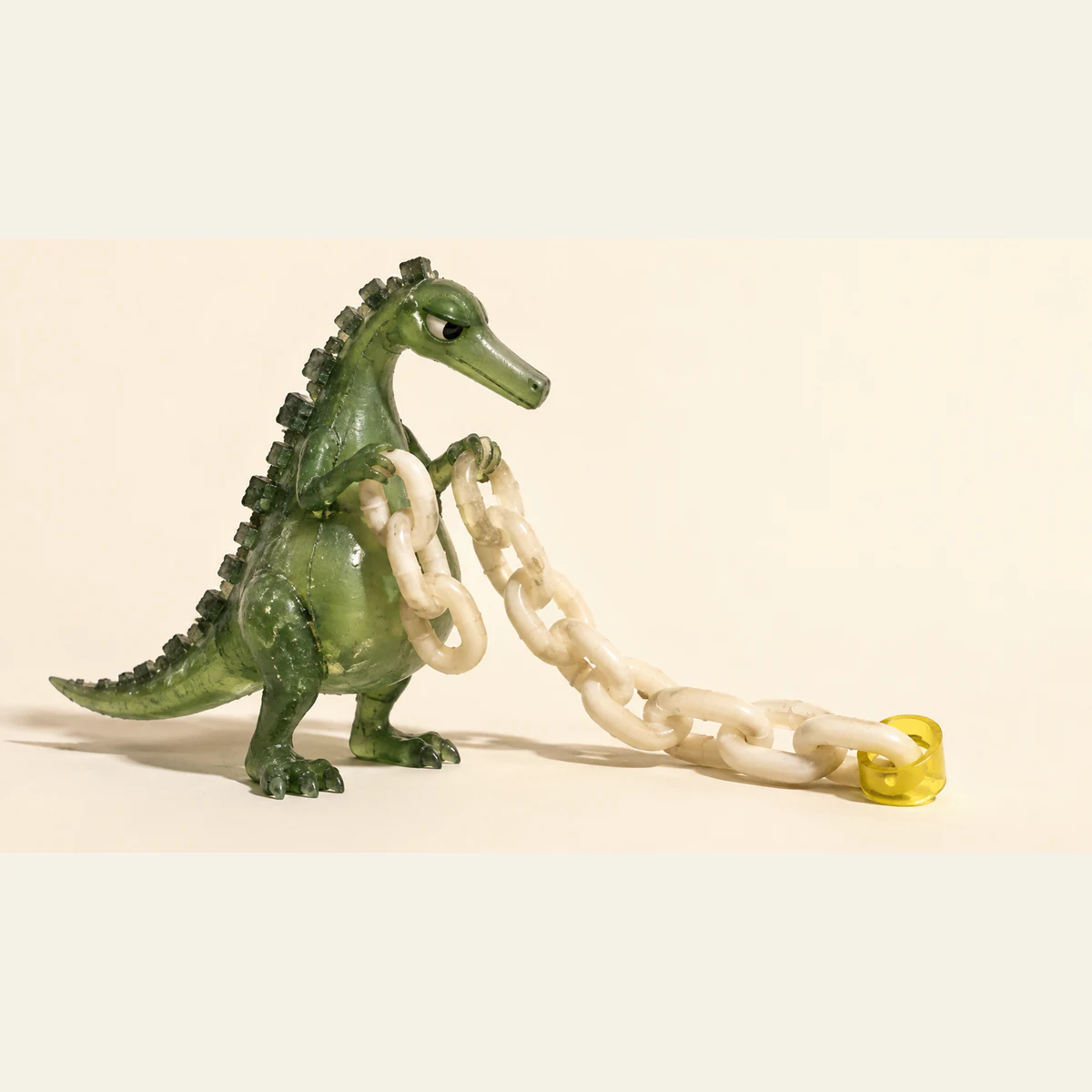 A translucent green resin dinosaur holds an ivory chain trailing across a warm cream surface. A yellow collar surrounds one end link, leaving the rest outside.