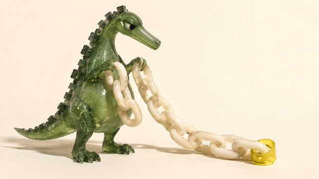 A translucent green resin dinosaur holds an ivory chain trailing across a warm cream surface. A yellow collar surrounds one end link, leaving the rest outside.