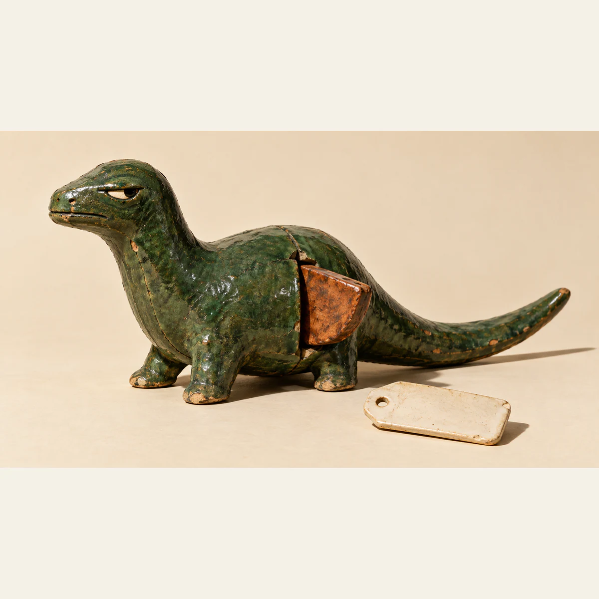 A scuffed forest-green ceramic dinosaur with a skeptical eye has a copper-colored insert protruding from an opening in its flank. A blank ivory tag lies detached beside it on a warm ivory surface, with hard shadows.