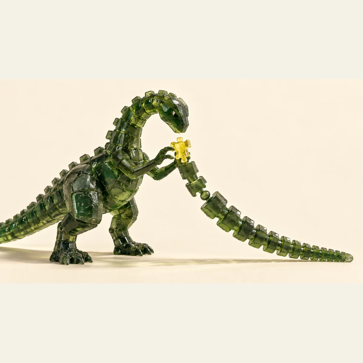 A translucent green resin dinosaur examines a yellow connector above a long segmented assembly on an ivory surface, with hard shadows and visible casting bubbles.