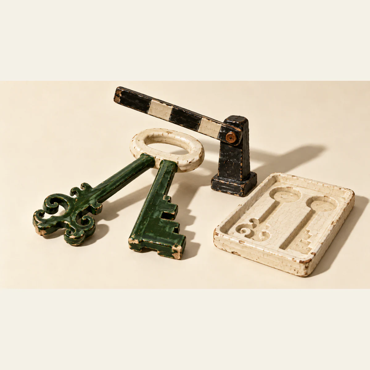 Two differently shaped forest-green ceramic key blades share an ivory handle beneath a raised black-and-ivory barrier. An empty tray with separate key-shaped recesses sits beside them. Chipped glaze and hard shadows texture the warm ivory scene.